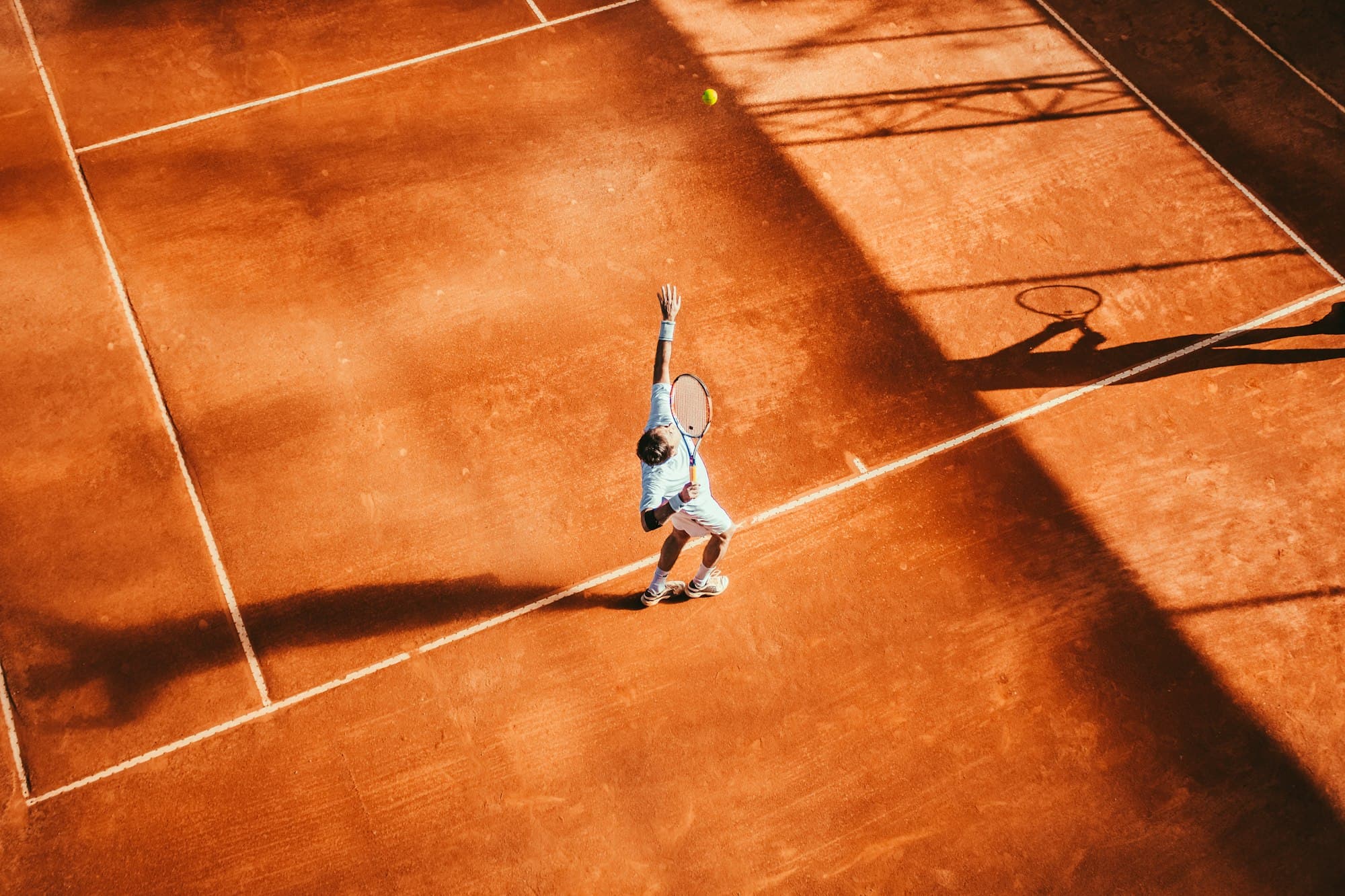 Overhead view of a serve on a clay court in late afternoon light.